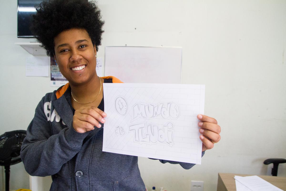 Moça segurando uma plaquinha escrito: O Mundo de Tludi